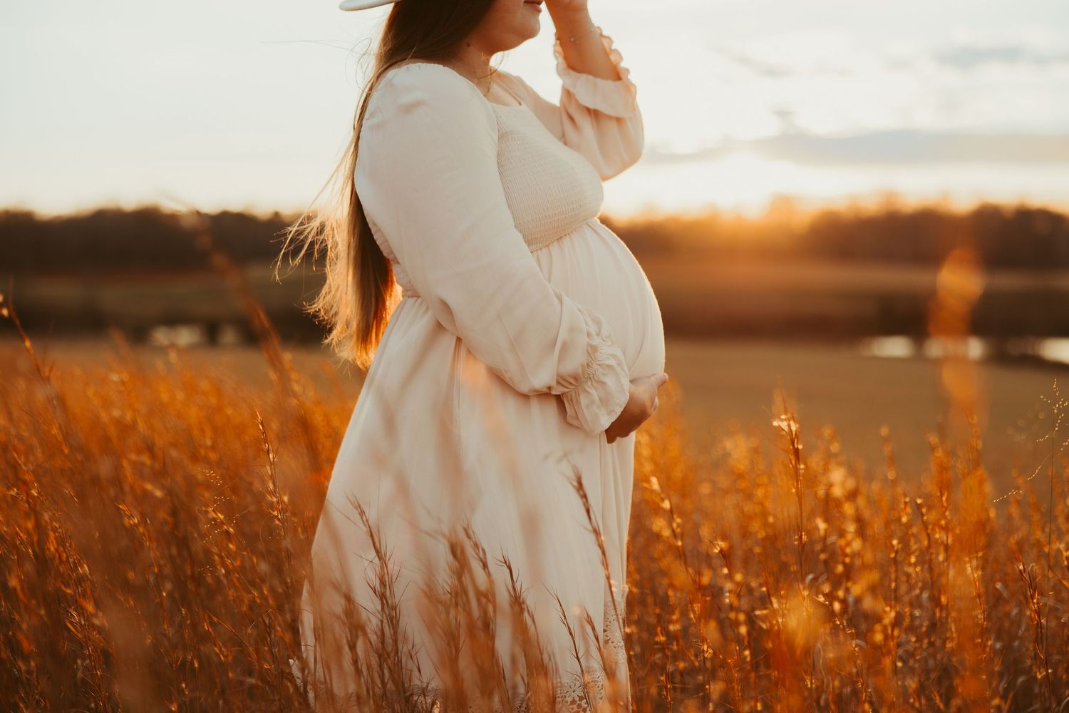 Pregnant woman in white dress cradling belly at golden sunset in field.