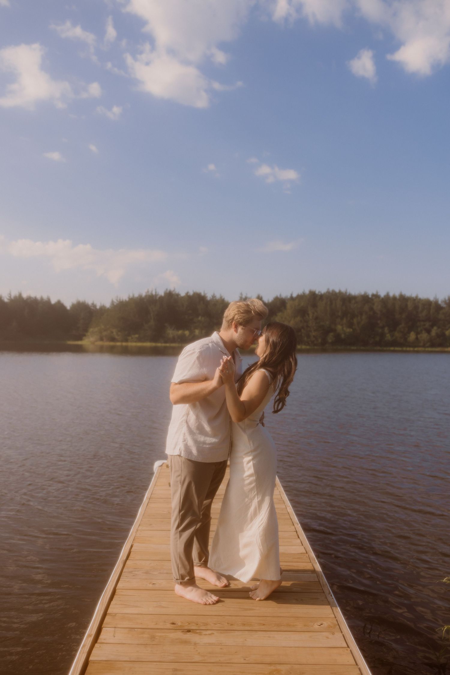 A couple in white outfits shares a romantic moment on a wooden dock overlooking a serene lake on a sunny day.
