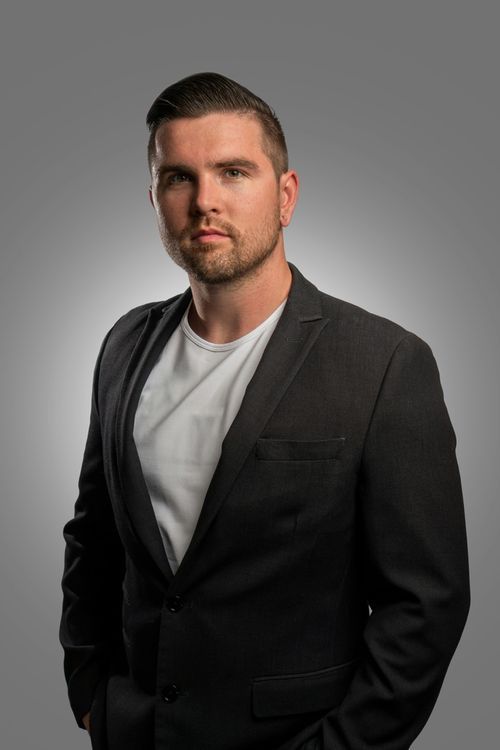Professional studio portrait or professional headshot of person in black blazer and white t-shirt against gray background.