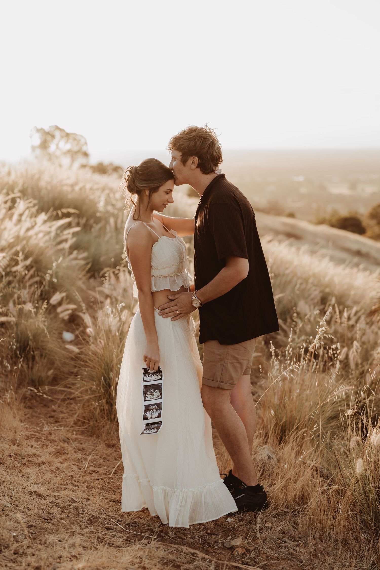 A couple embraces in a golden field during sunset while holding an ultrasound photo.