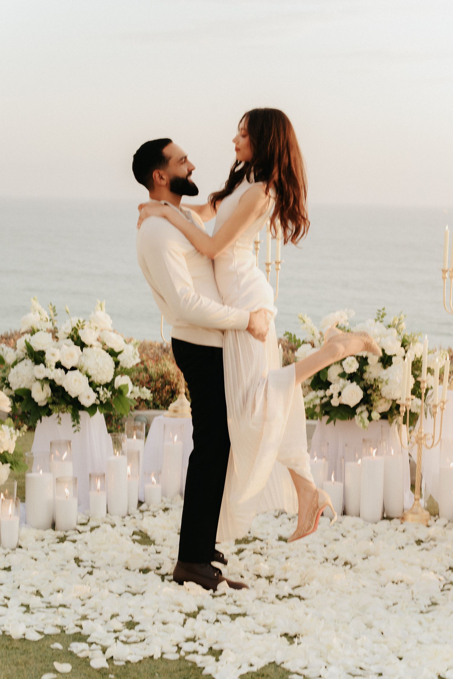 A romantic beach wedding scene with a couple dancing among white floral arrangements and candles at sunset.