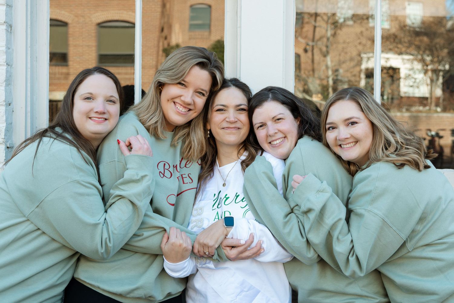 Bride surrounded by bridesmaids wearing matching sweatshirts — candid wedding celebration portrait