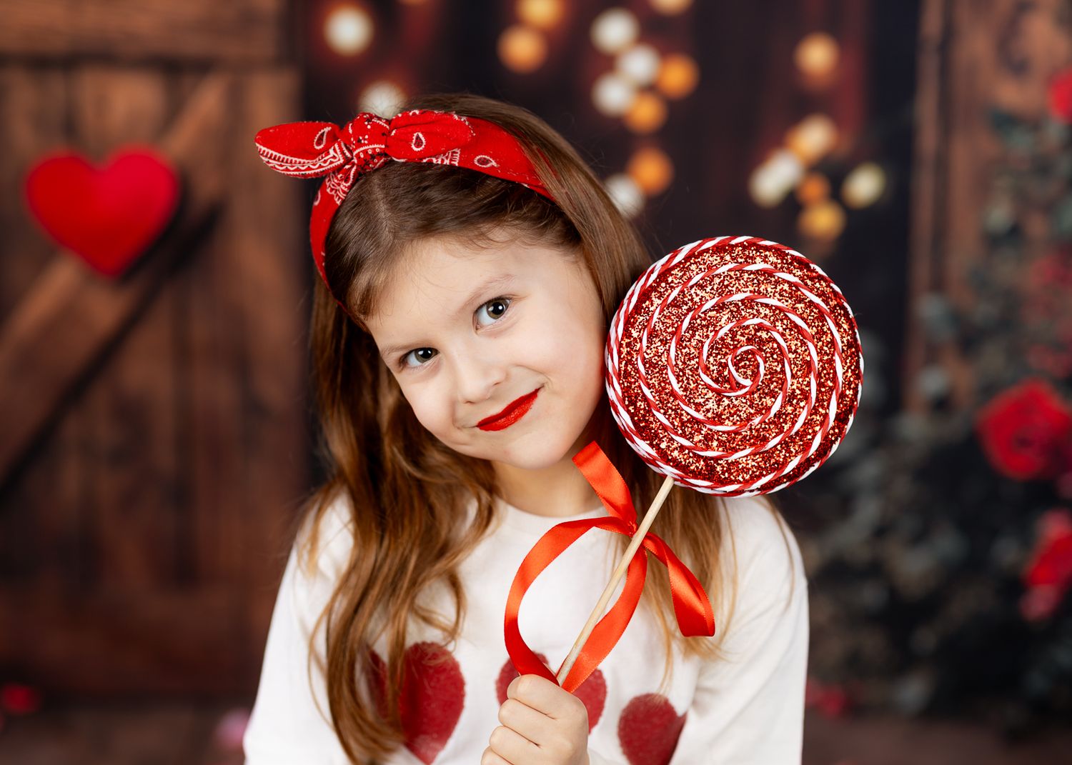 A child in a white dress holds a red lollipop while posing against a dark rustic background with red accents.