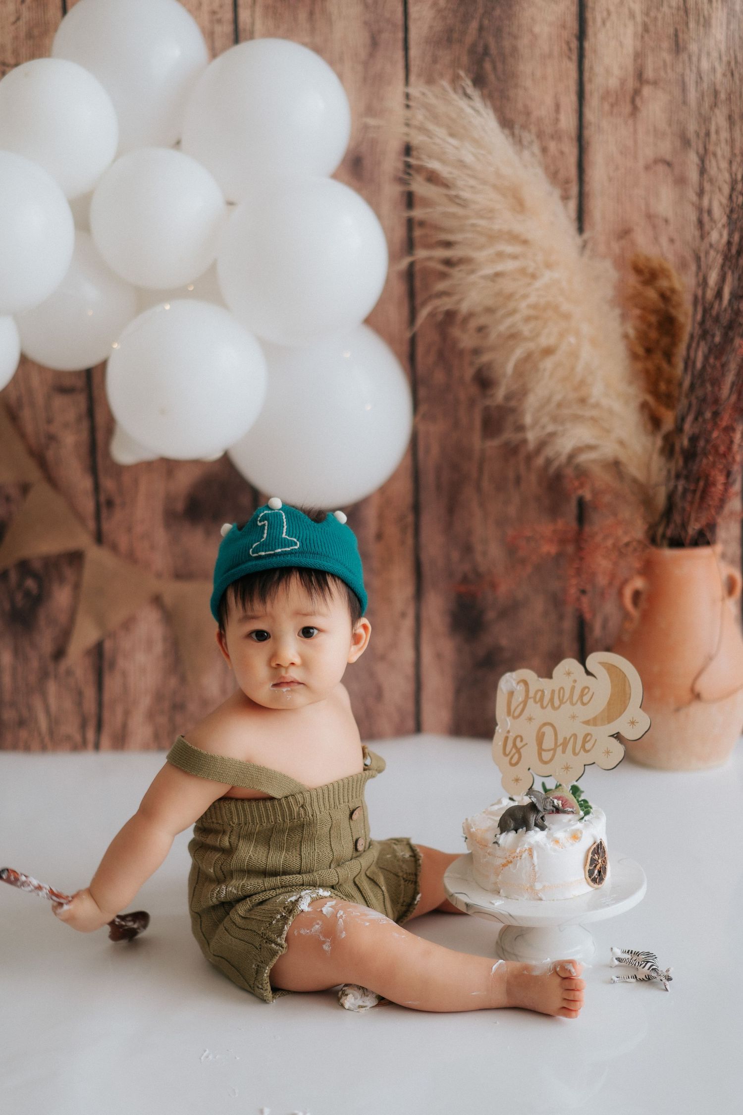 A baby cake smash photo session with white balloons, pampas grass, and rustic wood backdrop for first birthday celebration.