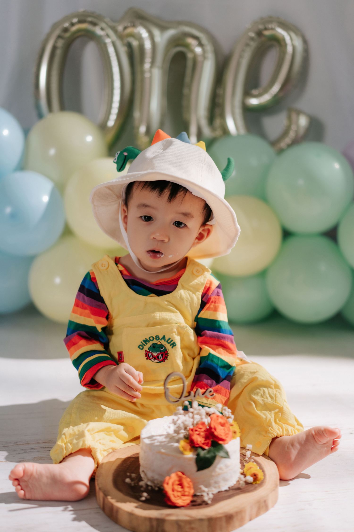 Baby in white outfit plays with colorful pastel balloons and smash cake during first birthday photoshoot.
