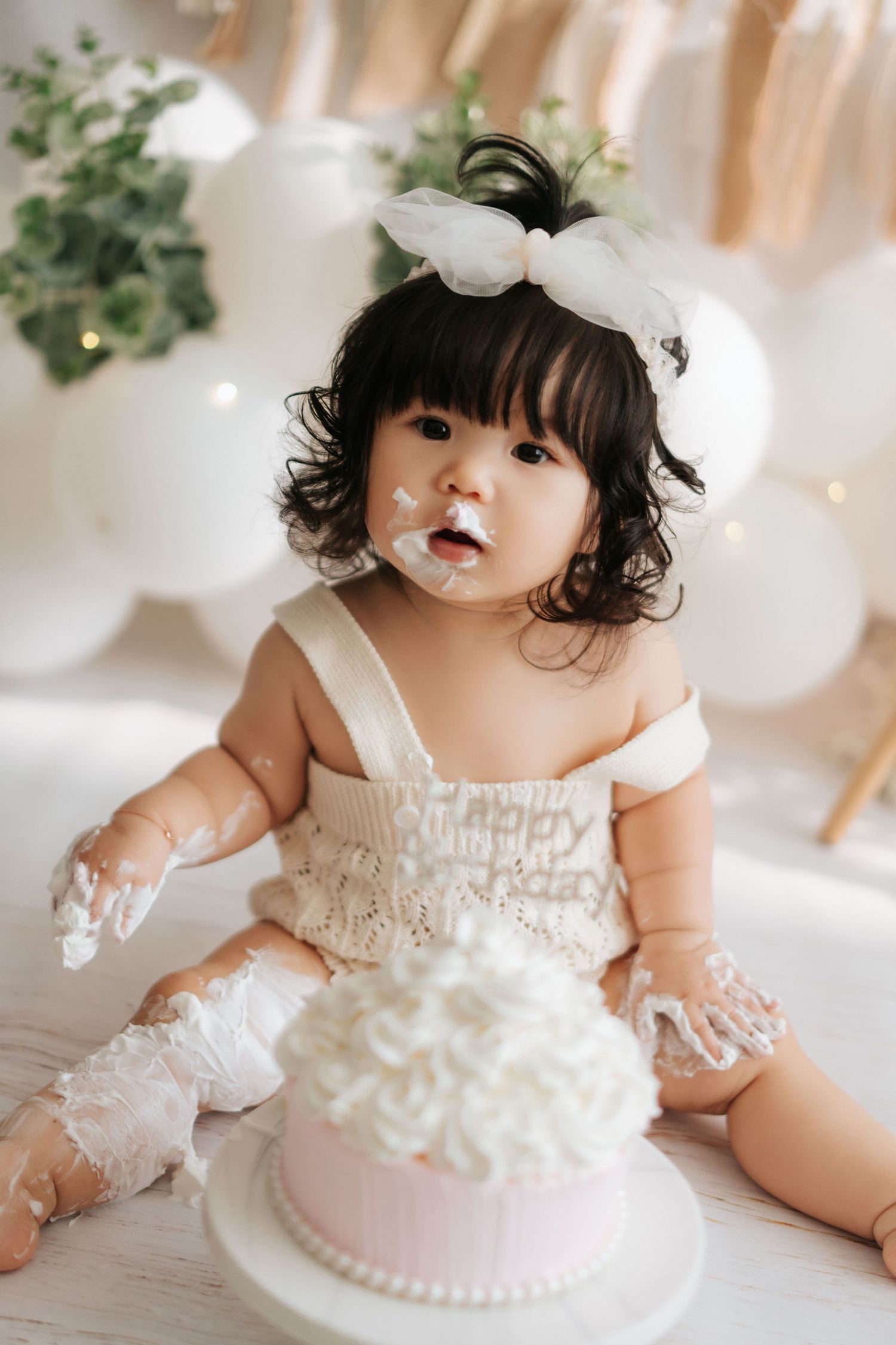 A cake smash photo session featuring a baby in a white dress surrounded by balloons and decorative elements.