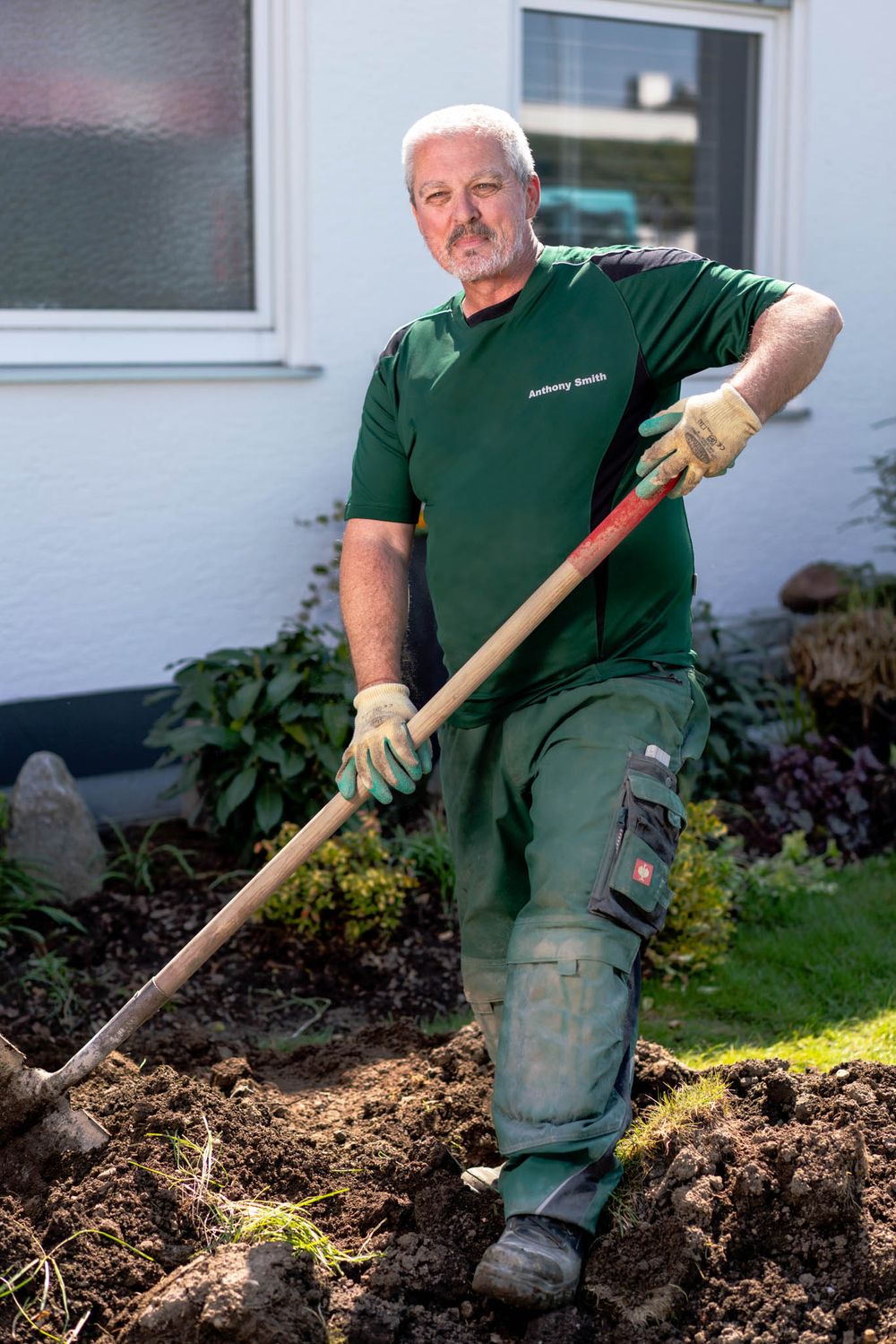 Landscaper digging and preparing soil in a garden bed next to a white house exterior.