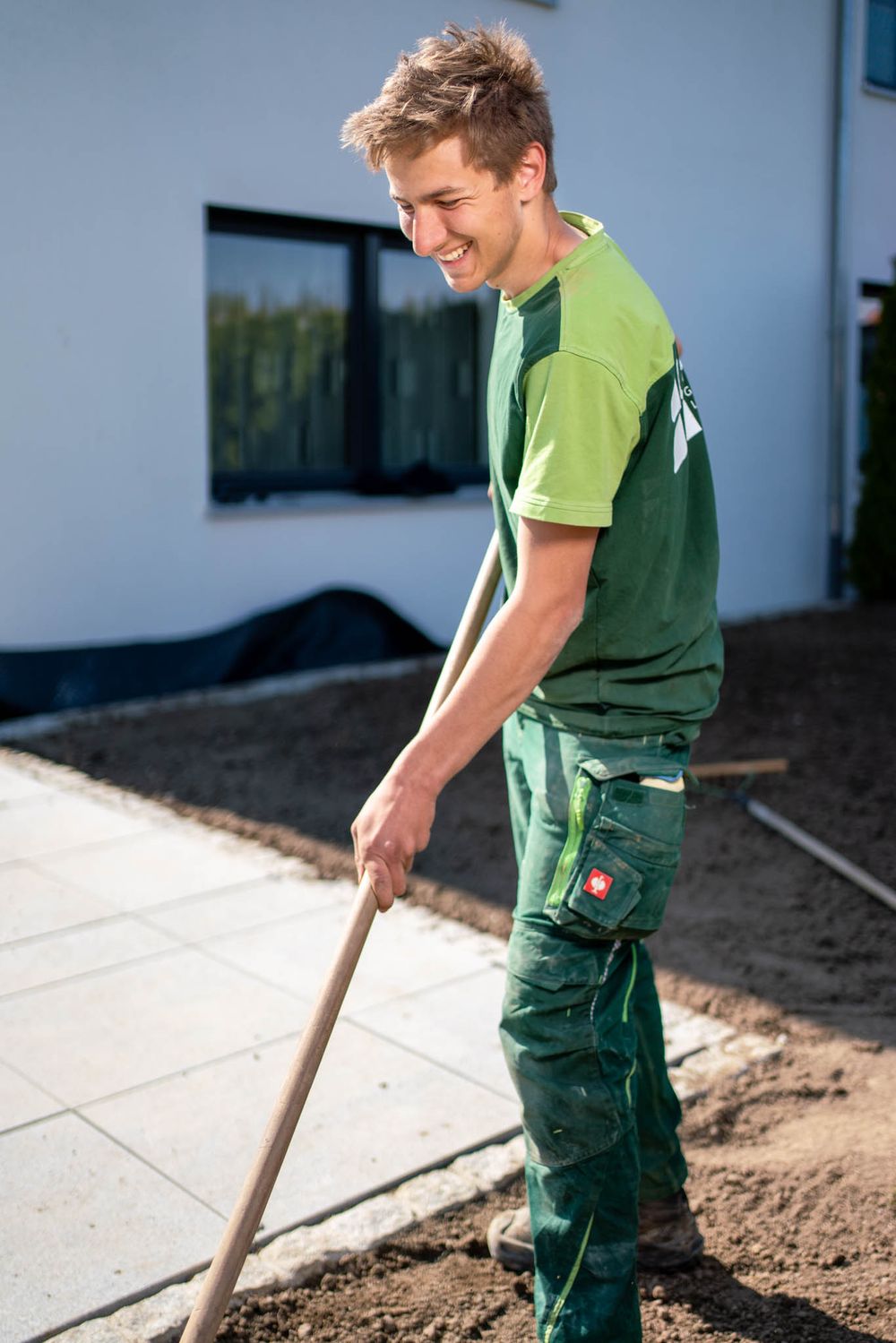 Landscaper in green work uniform smiles while using a rake on dirt pathway during outdoor maintenance work.