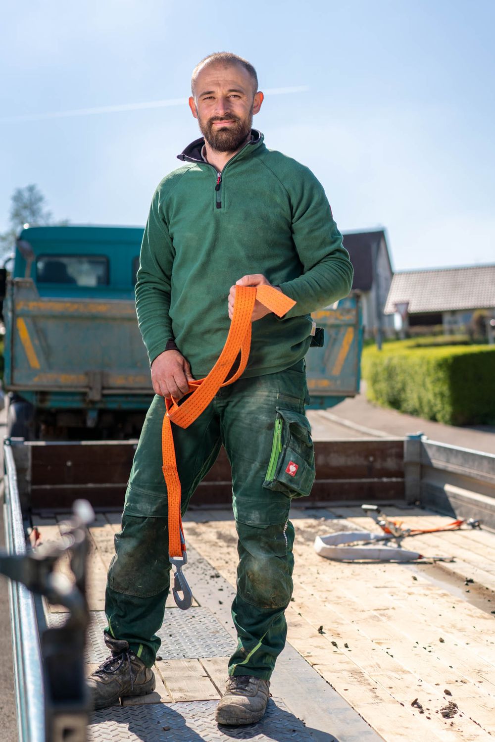 Person in green workwear and orange gloves standing on construction site with dump truck in background.