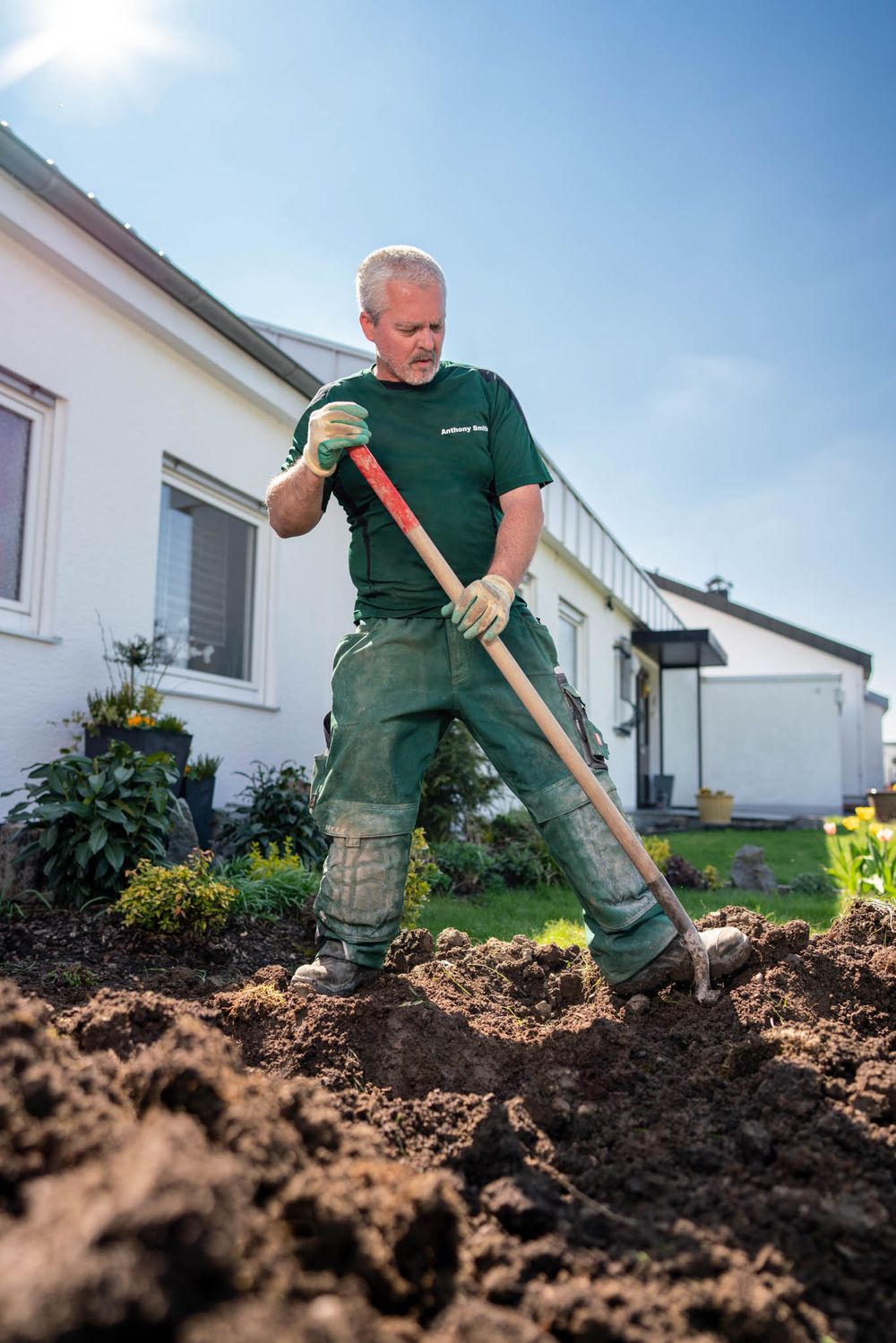 Gardener in green uniform using a spade to dig and turn soil in a residential garden bed.