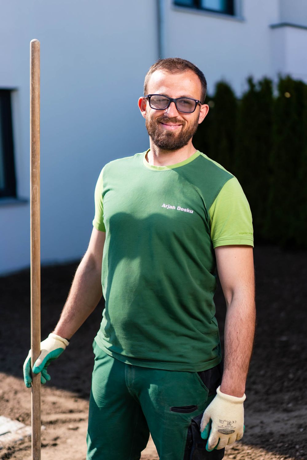 A gardener in green work attire smiles while working outdoors with wooden stakes in the ground.