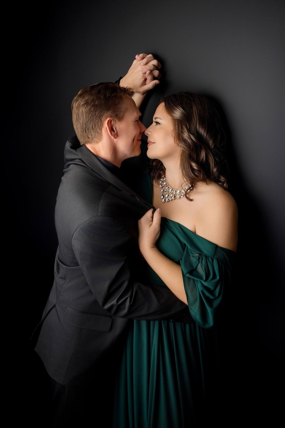 Romantic couple embrace against dark backdrop, with woman in green dress and statement necklace Knoxville TN couple photography session.