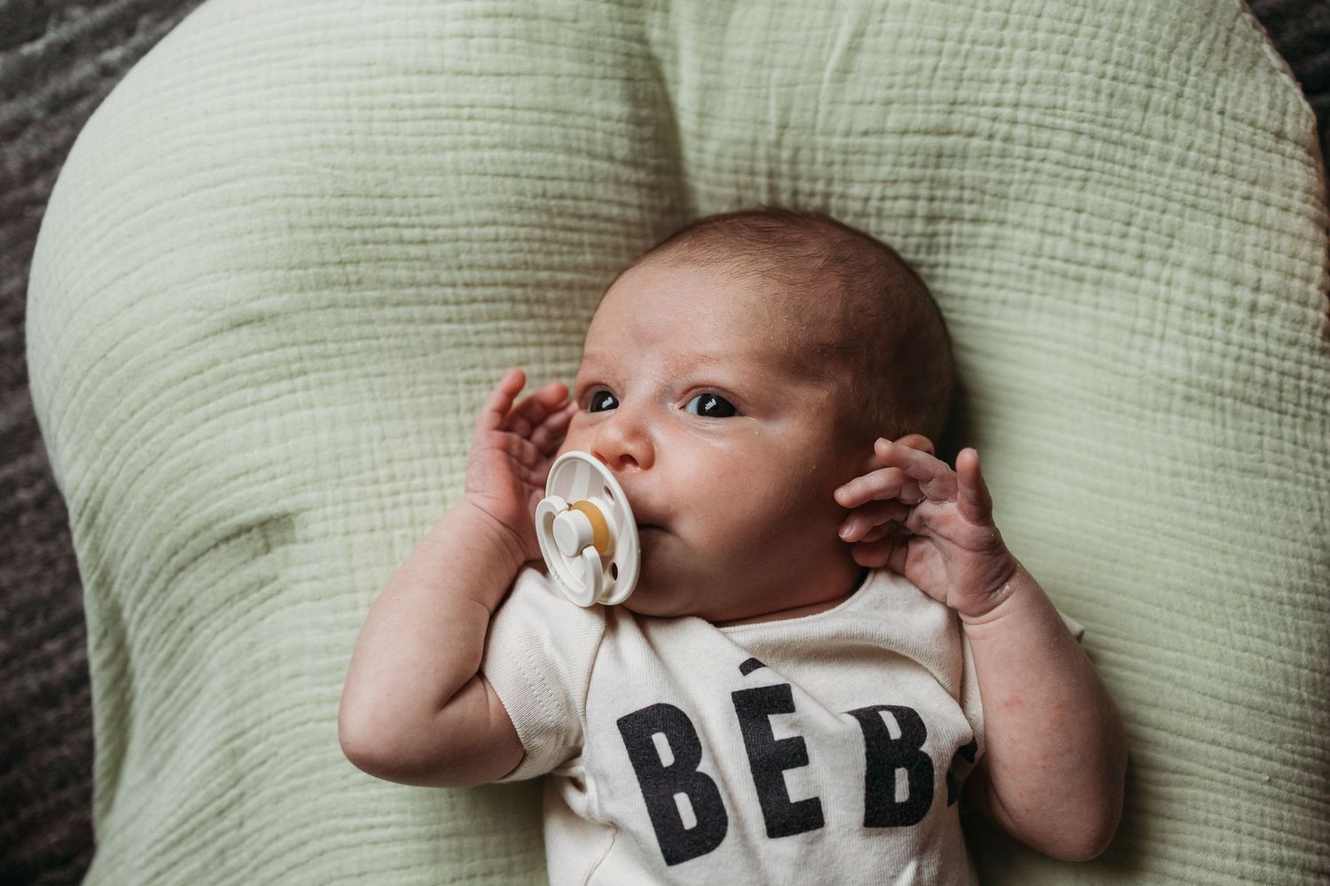 Baby lying on mint green cushion wearing a 'BEBE' onesie with a pacifier in their mouth.