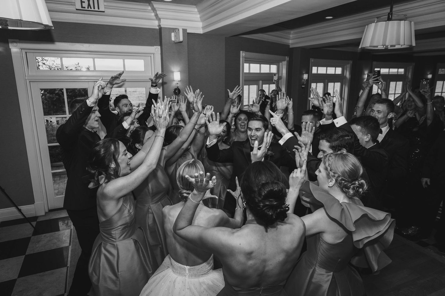 Wedding guests dance enthusiastically with raised arms on a crowded dance floor at an indoor reception venue.