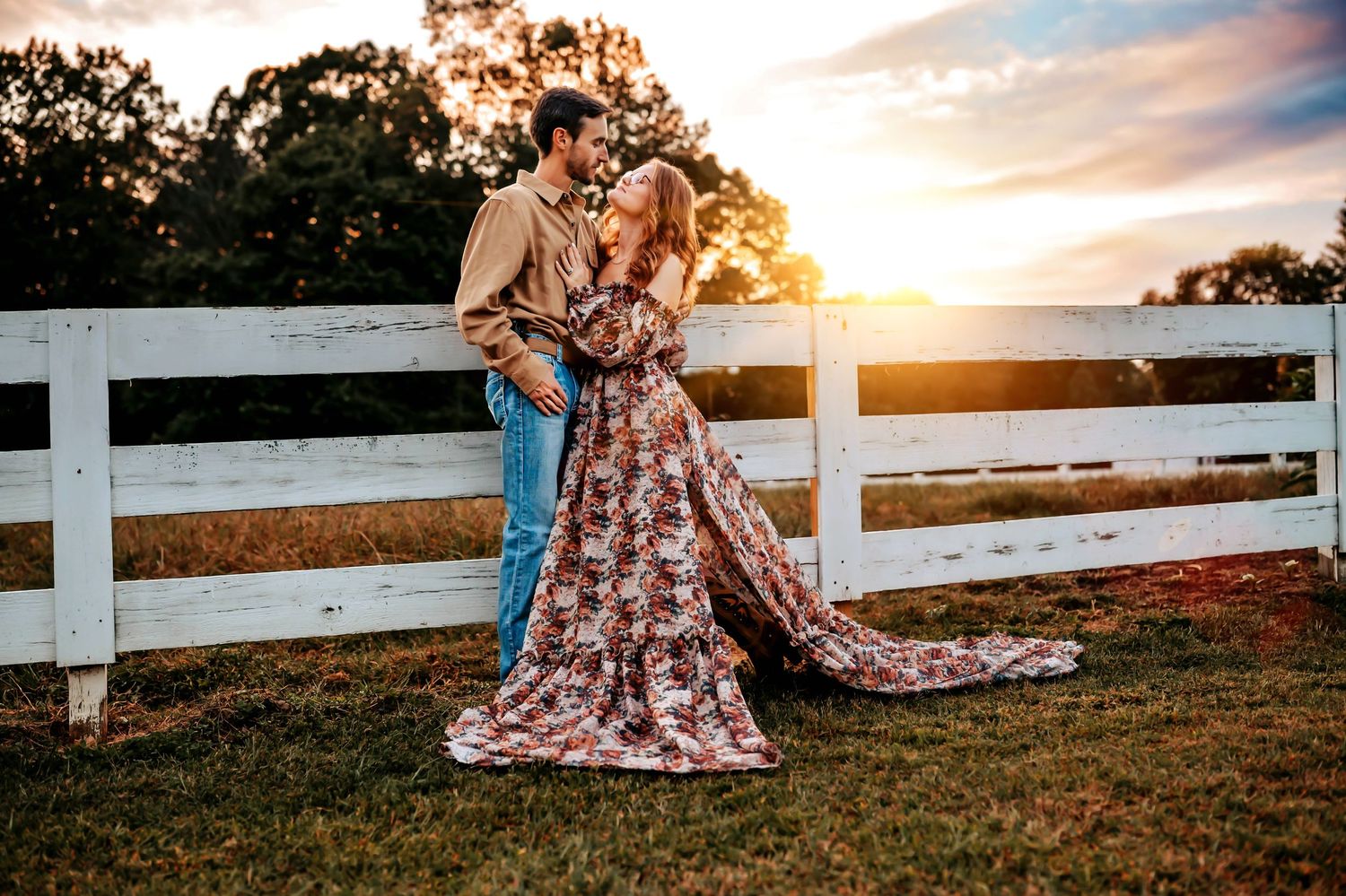 A romantic couple embraces at sunset near a white fence on a grassy field during a golden hour photoshoot.