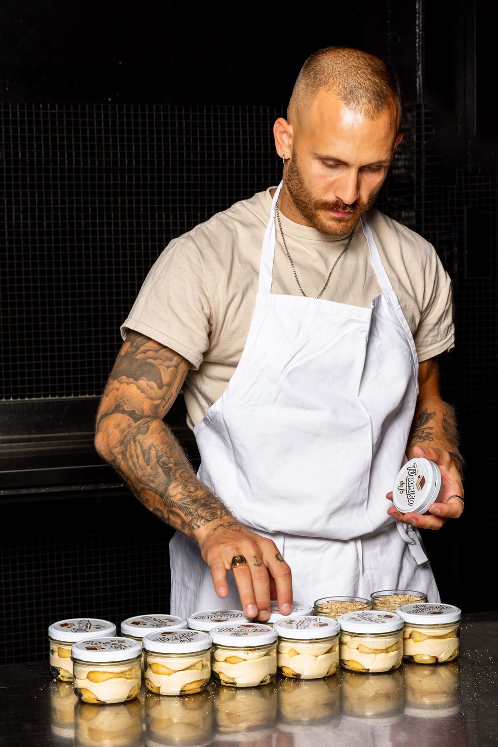 Chef in white apron arranging rows of desserts in glass jars on a dark counter.
