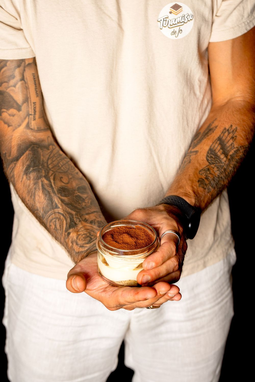 Tattooed hands holding small white desserts against a black background.