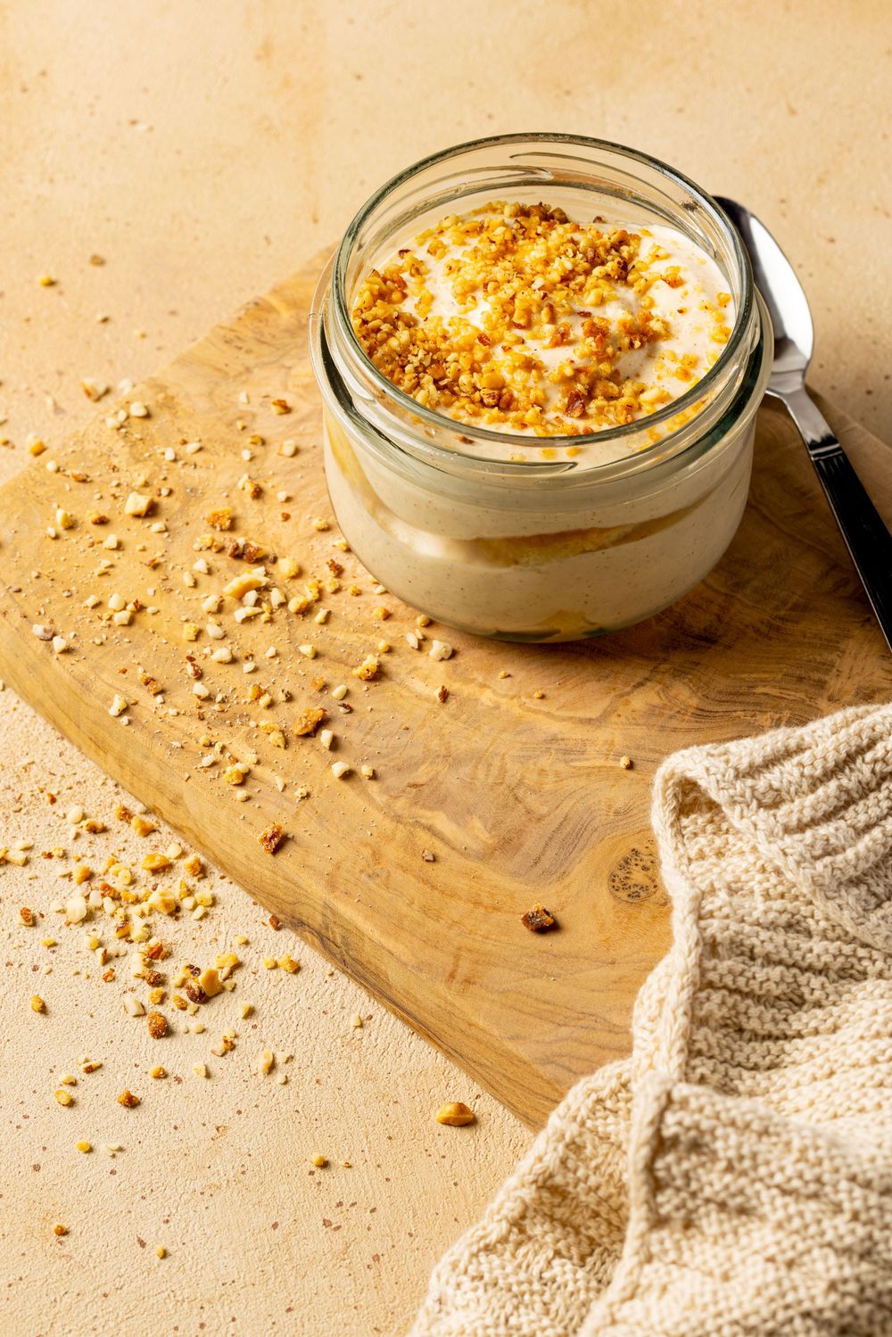 Creamy dessert in glass jar topped with crushed nuts and crumbs on wooden board with knitted cloth.