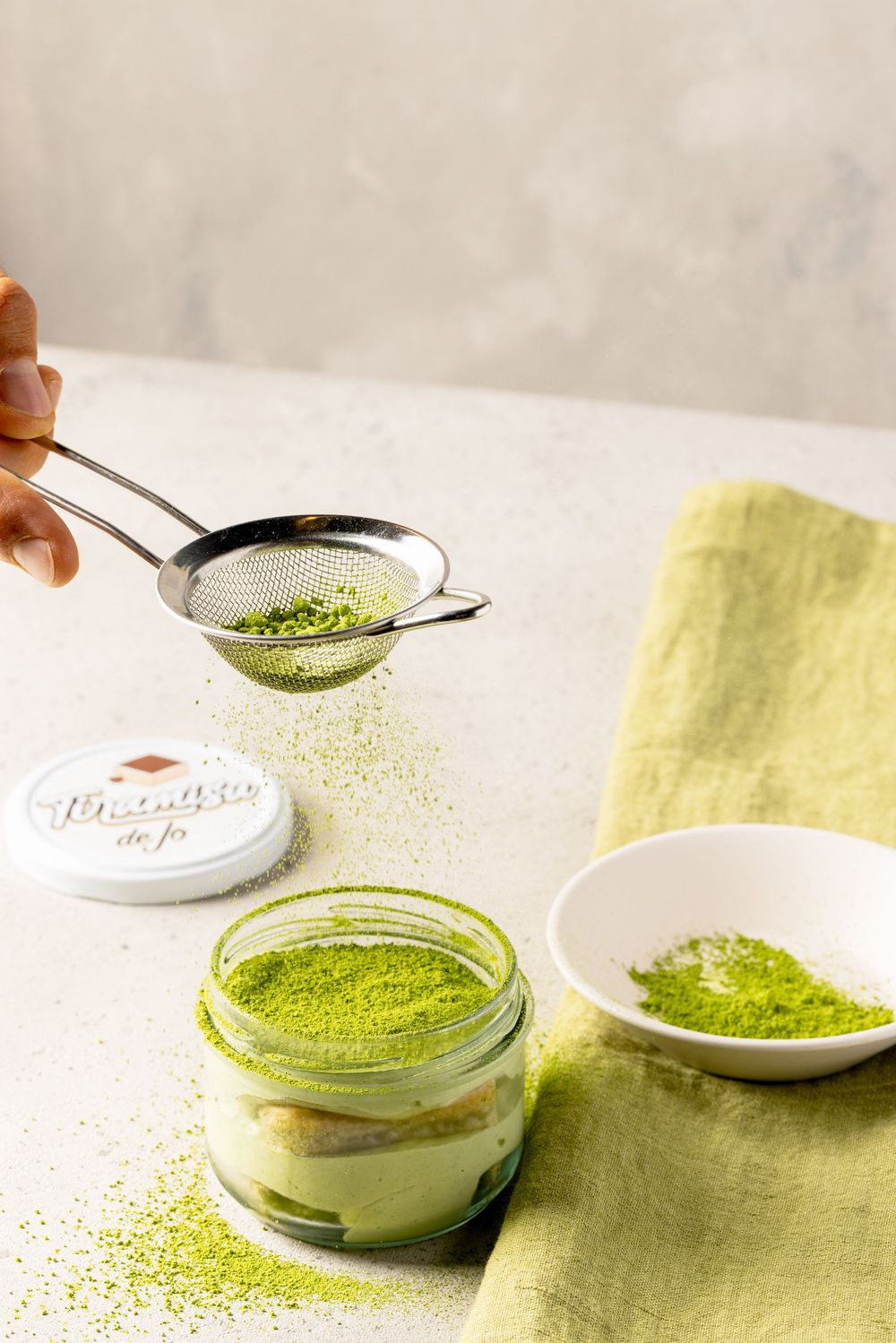 Sprinkling bright green matcha powder onto a white cream dessert in glass jars on a light surface.