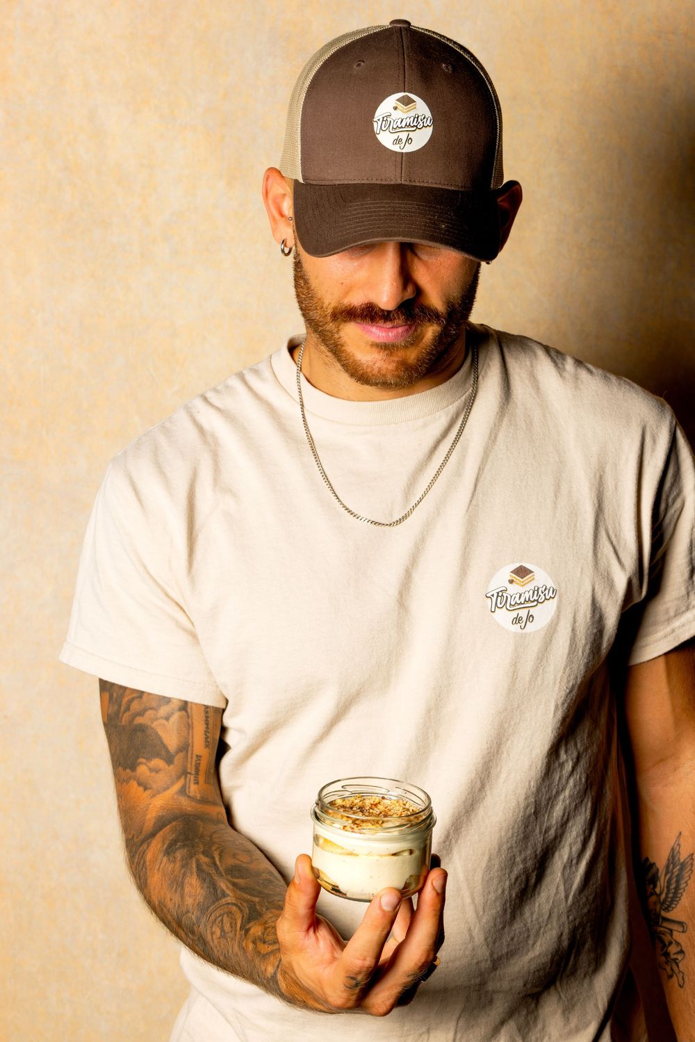 Person wearing brown cap and white t-shirt holding jar of dessert against beige wall.