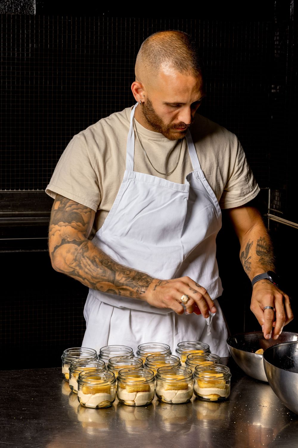 Chef with tattooed arms carefully arranges and prepares desserts in glass jars at a restaurant counter.