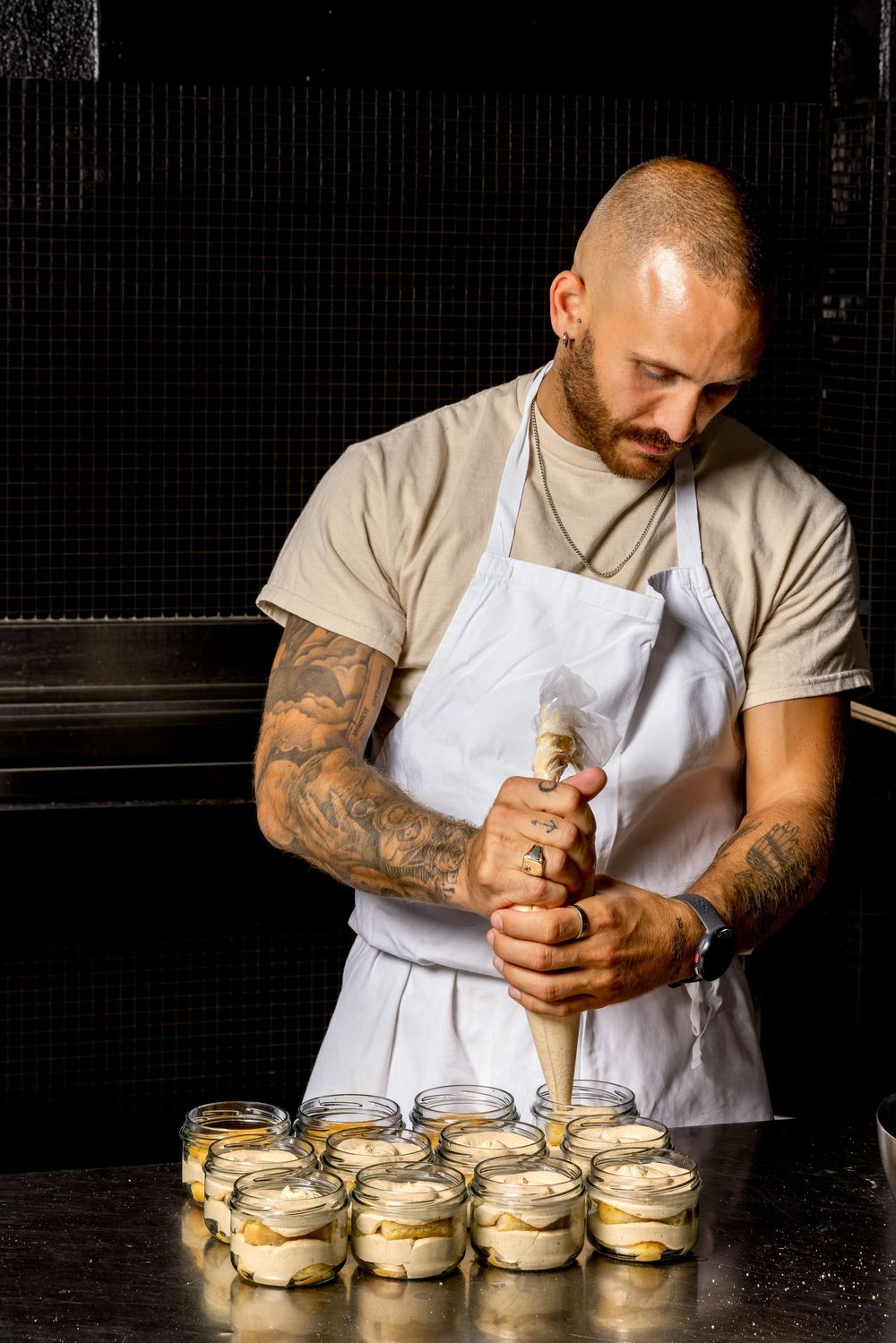 Chef in white apron carefully sealing glass jars filled with creamy dessert against dark background.