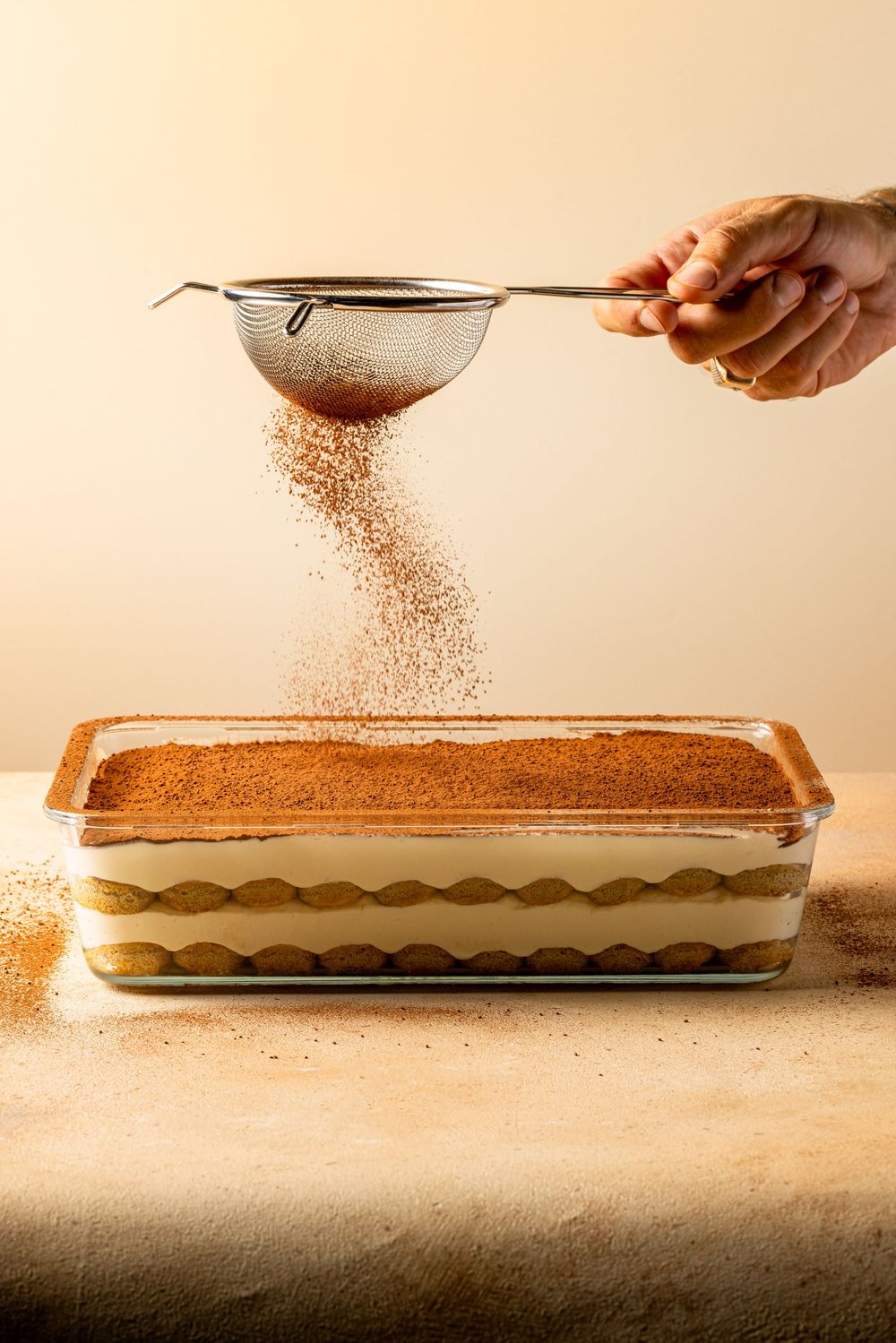 Hands sprinkling cocoa powder over a rectangular baking dish in a sequence of preparation steps.