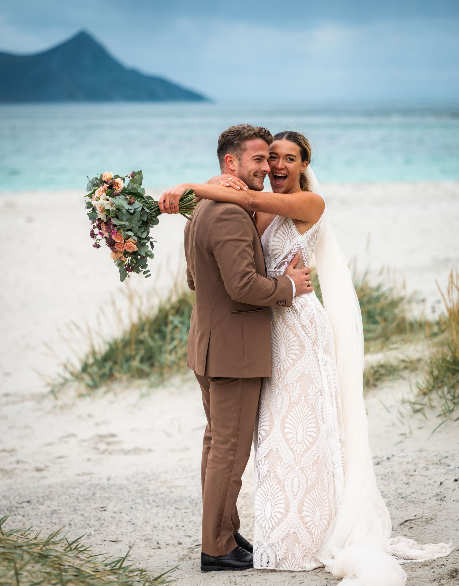 A couple shares a romantic moment on a beach with ocean waves and sand dunes in the background.