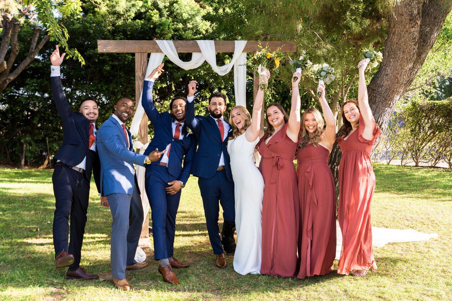 Wedding party in blue and dusty rose attire poses together outdoors on a sunny day.