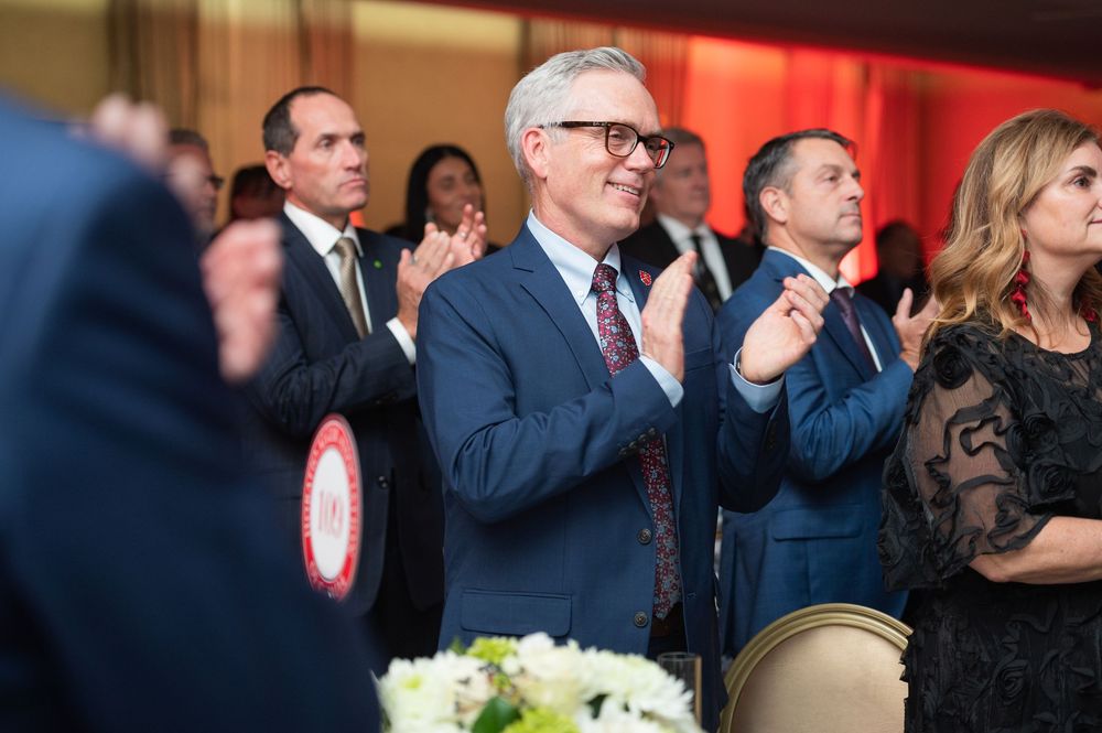 Audience members in formal attire applaud during an event while seated at decorated tables in a red-lit venue.