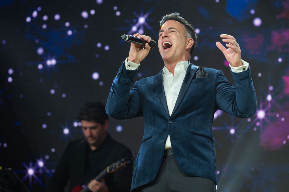 Performers in dark suits gesture dramatically on stage with colorful spotlights and sparkles in the background.