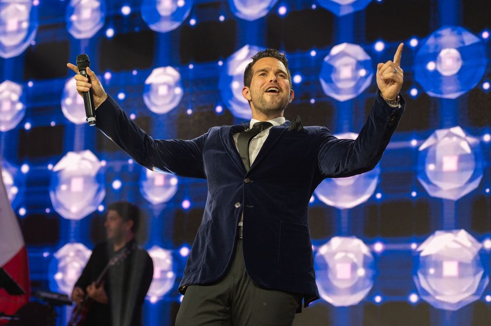 A performer in a dark suit raises their arms triumphantly on a stage with glowing blue lights in the background.