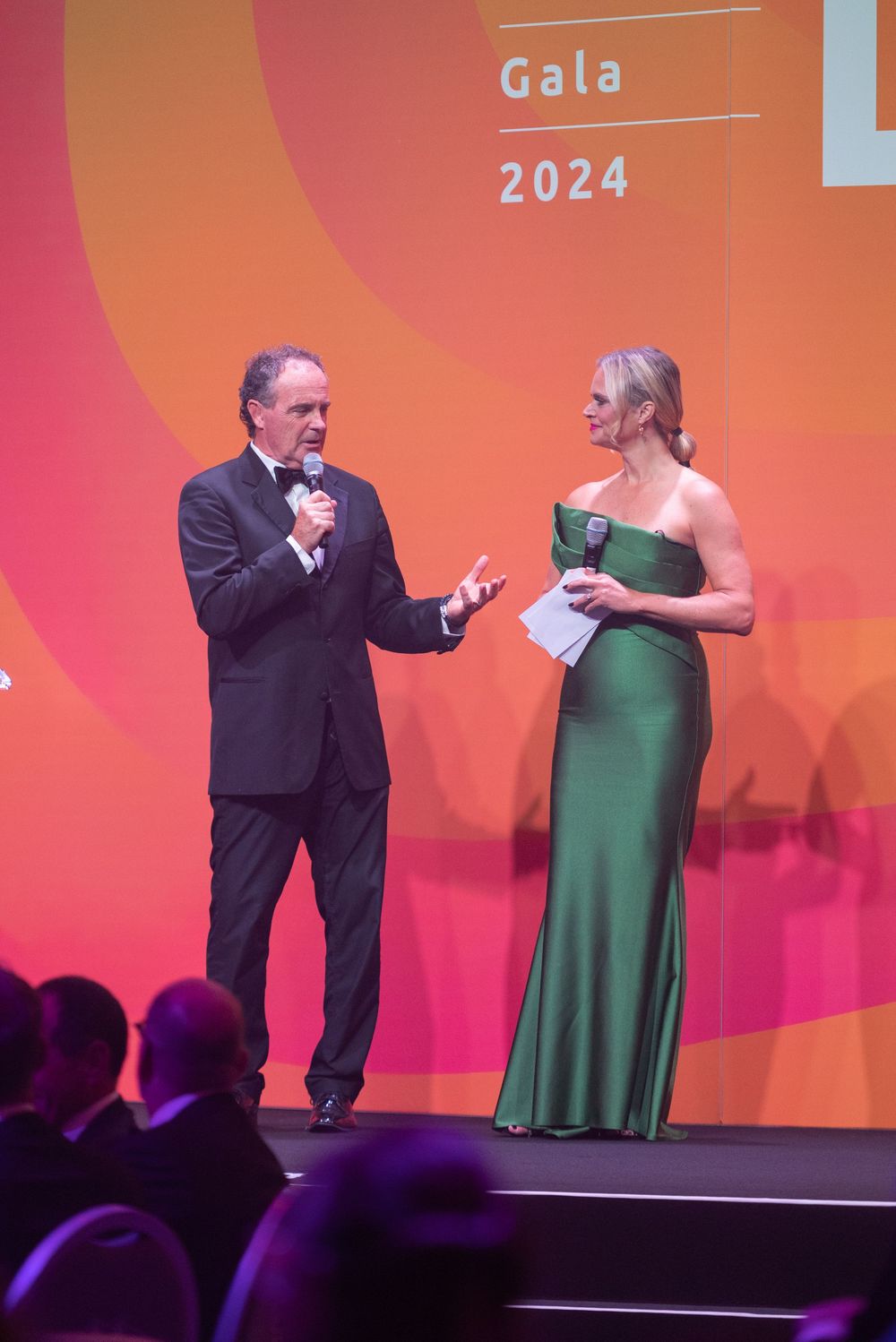 Two performers interact on stage during a theatrical performance against an orange backdrop.