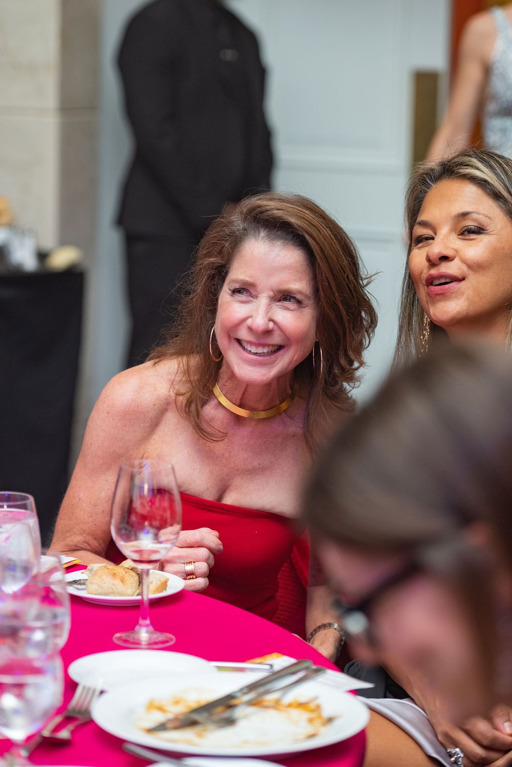 A person in a red dress laughs while dining at a restaurant table with plates of food.