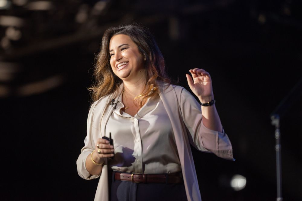 A speaker on stage holds a beverage while gesturing enthusiastically during a presentation in a dark auditorium.