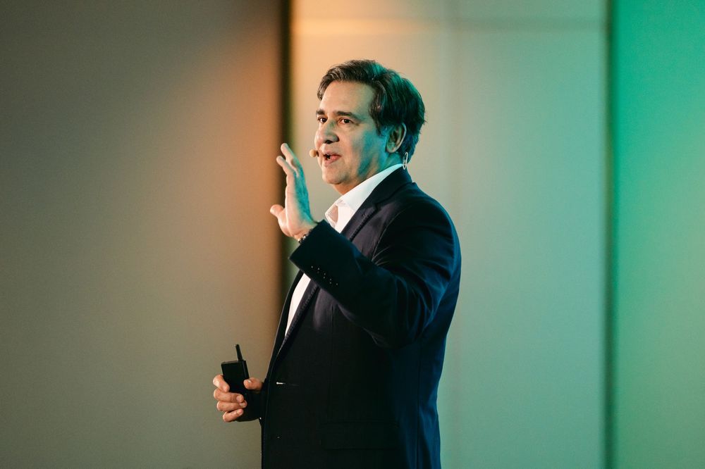 A person in a dark suit gestures animatedly while speaking against a bright turquoise background.