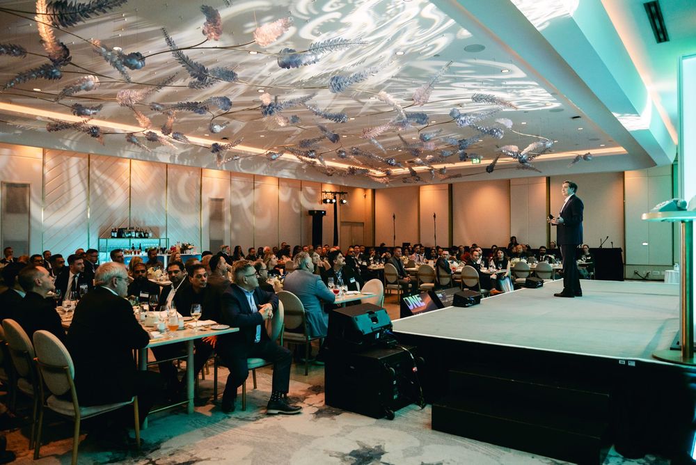 A large conference room filled with attendees watching a speaker on stage under decorative ceiling lighting.