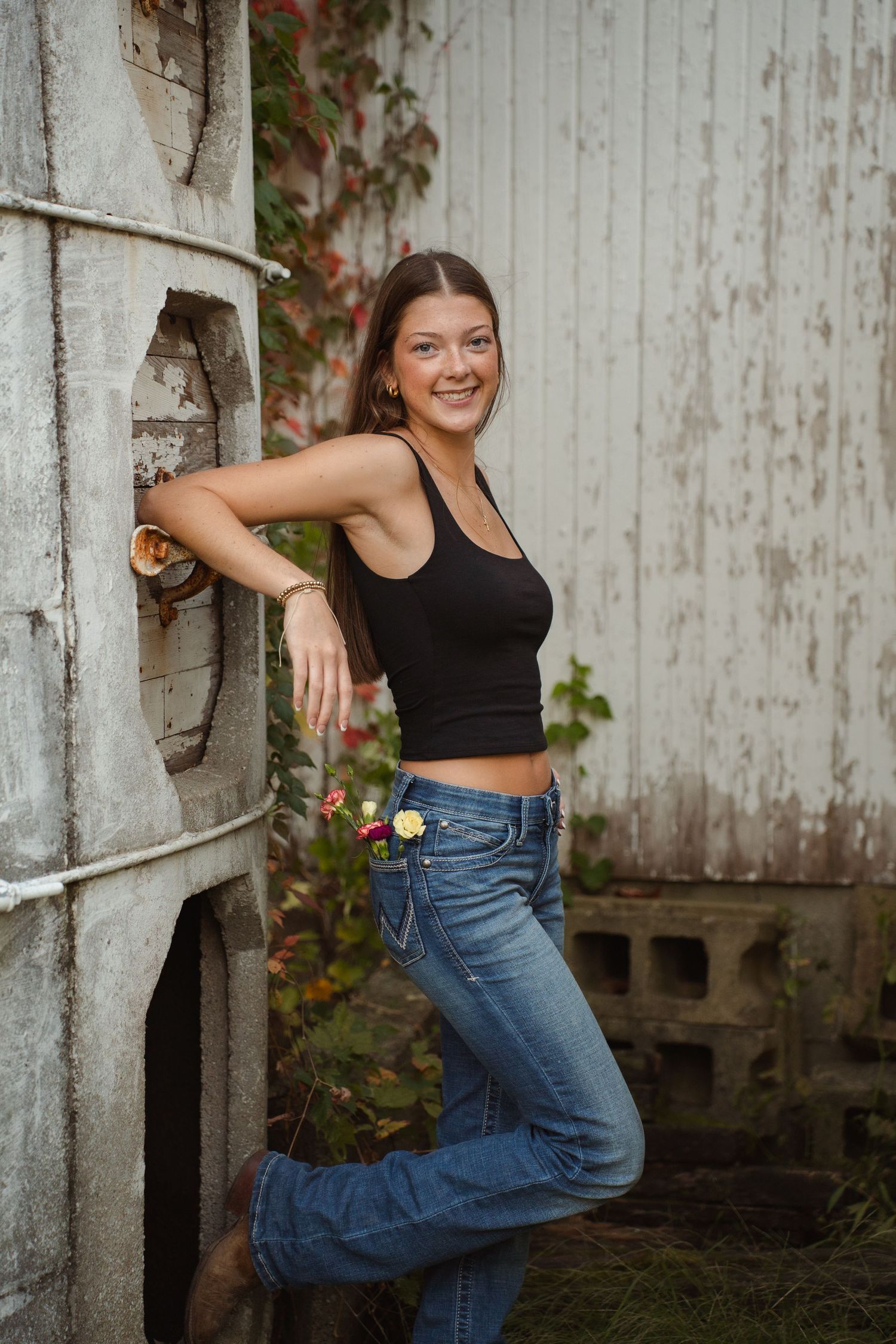Person posing casually against rustic white silo wall wearing black tank top and jeans.