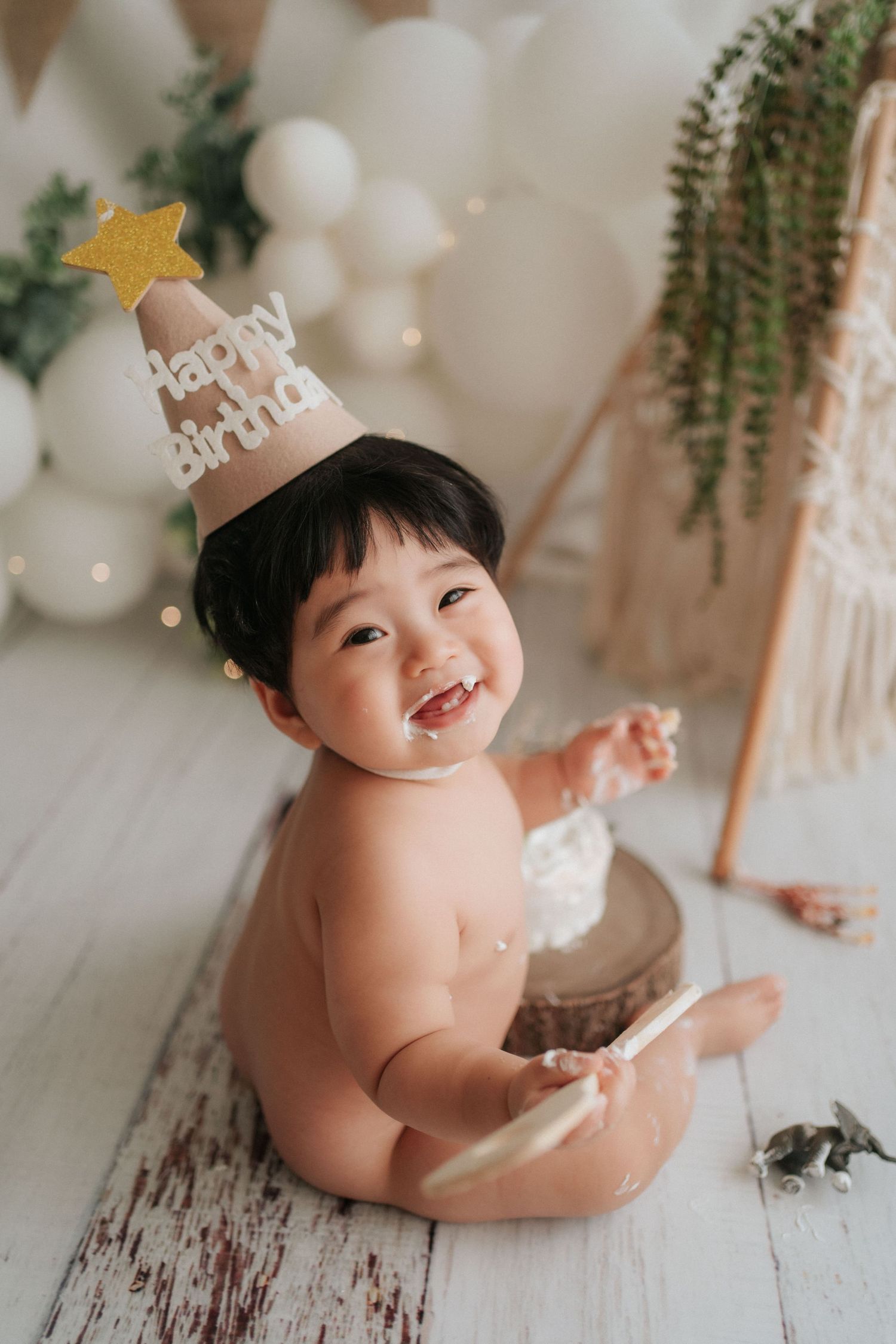 A baby wearing a birthday crown sits on a white floor surrounded by balloons and hanging greenery decorations.