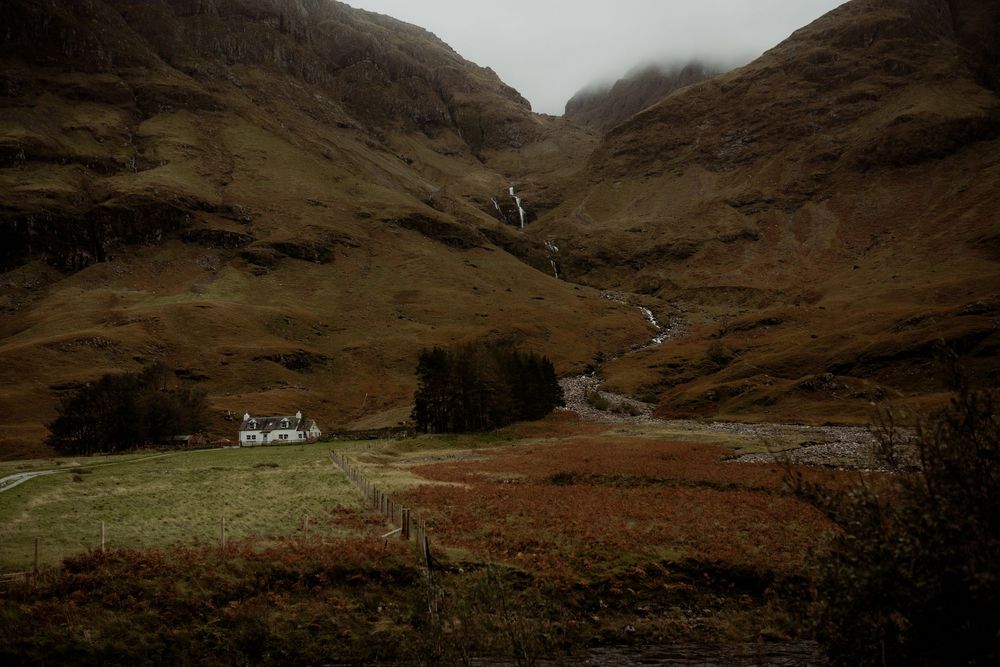 A remote white cottage nestles at the foot of a dramatic Scottish glen with cascading waterfalls on the mountainside.