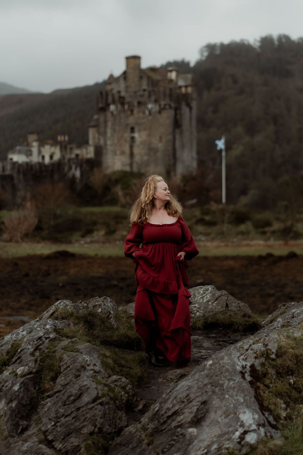 A figure in a flowing red dress stands on rocky coastal terrain with a castle visible in the misty background.