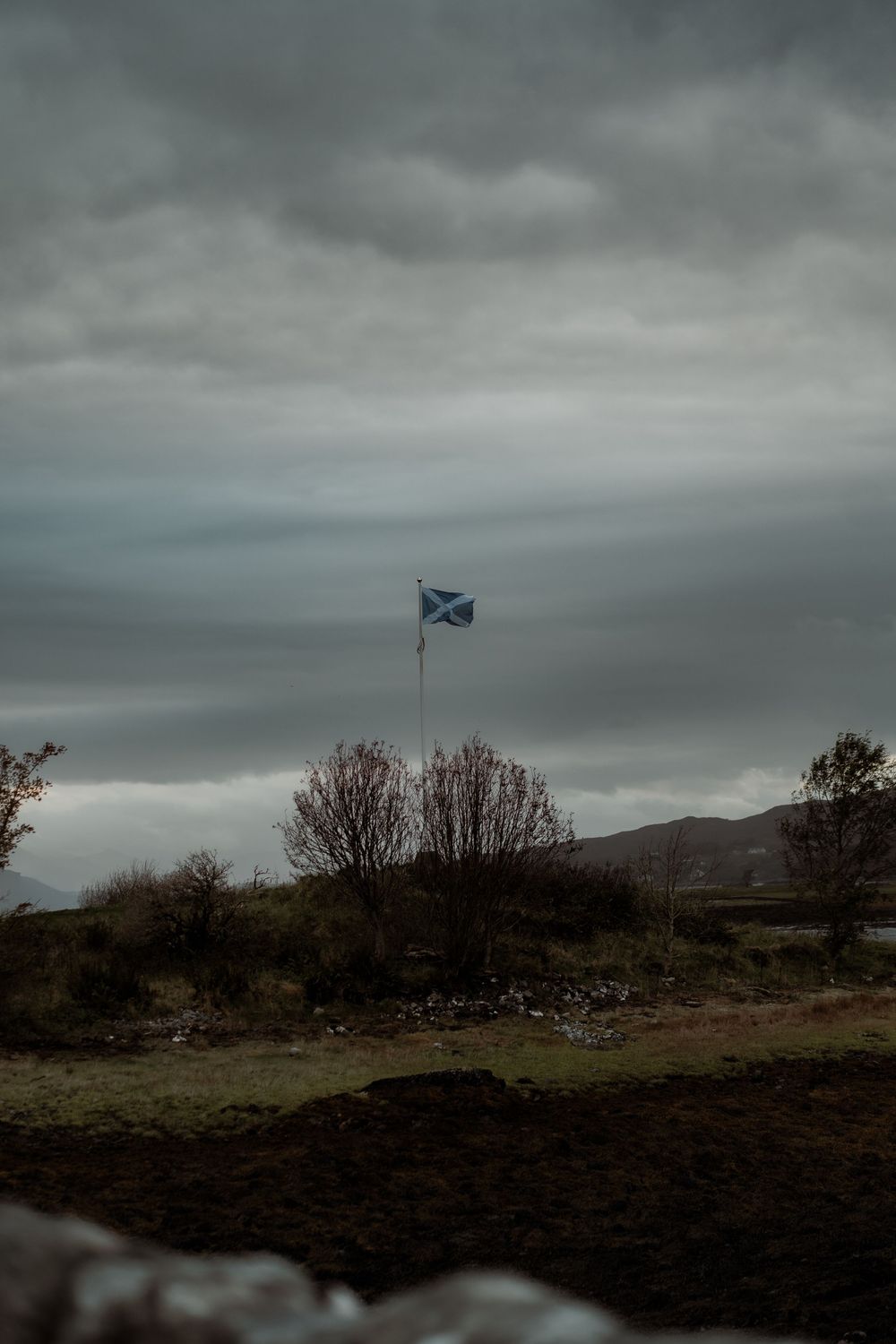 A Scottish flag waves against a moody, overcast sky above rugged highland terrain under dark storm clouds.