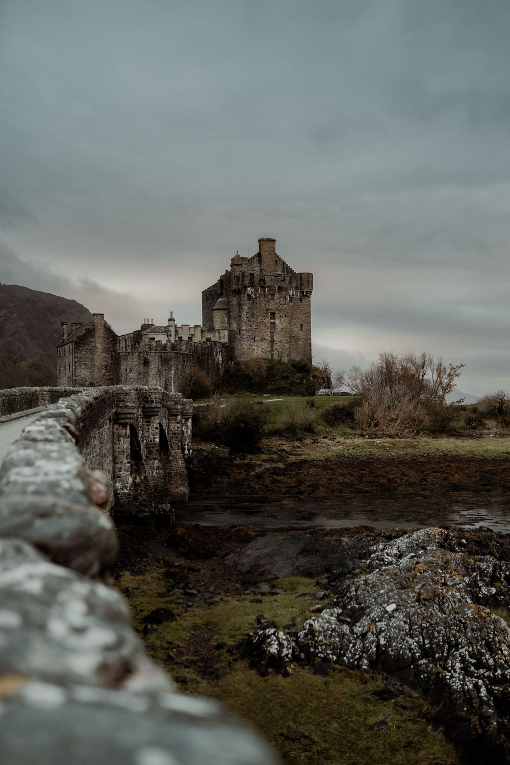 Moody view of Eilean Donan Castle in Scotland on an overcast day with dramatic stormy skies and rocky terrain.