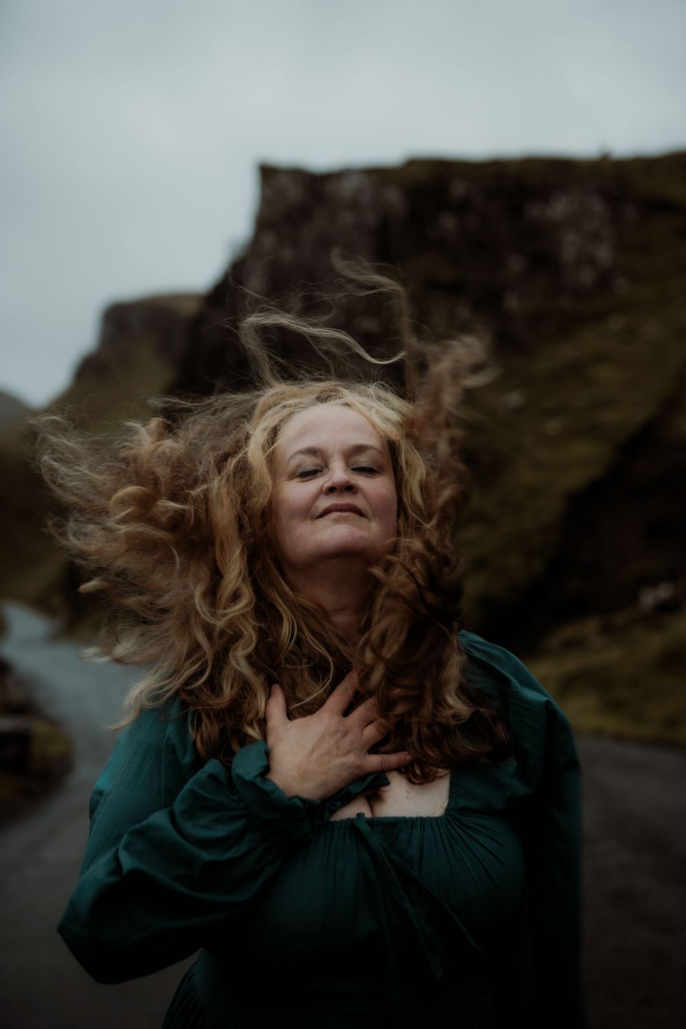 Hair blowing dramatically in the wind against a moody coastal backdrop with stormy skies.