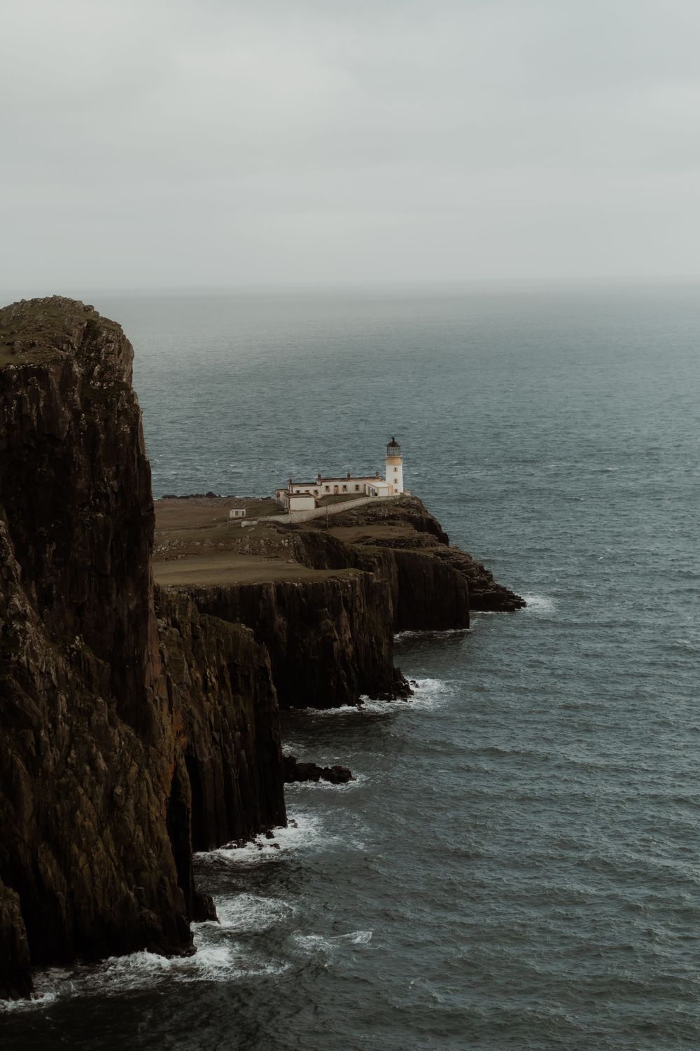 Dramatic coastal landscape with white lighthouse perched on rugged cliffs above the misty ocean at Neist Point, Scotland.