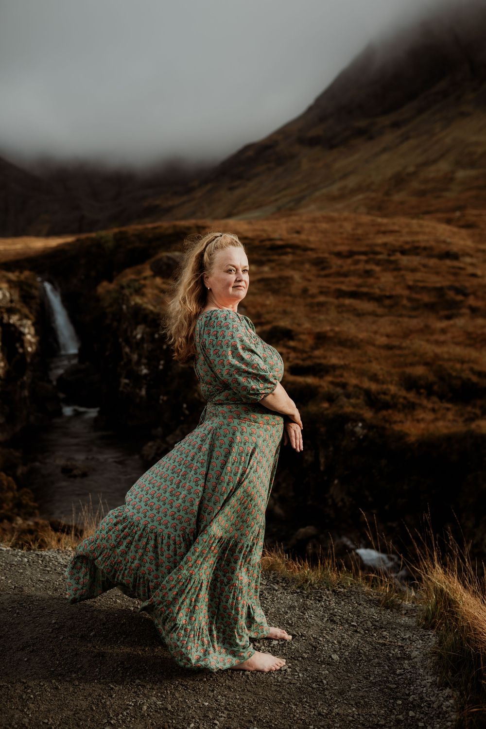 Person in a flowing green dress twirls against a scenic backdrop of Scottish highlands with a cascading waterfall.