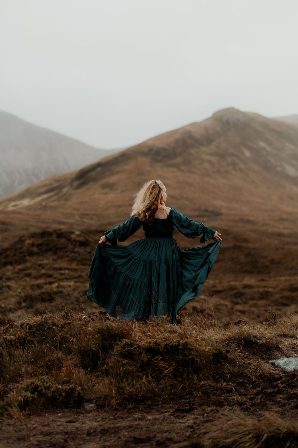 A dark teal dress flows gracefully in the wind against the backdrop of rugged Scottish highlands on a moody day.