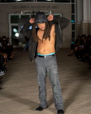Model presents urban streetwear look with dark jacket and cargo pants on runway.