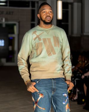 Model walks runway in light beige sweatshirt and distressed denim jeans during fashion show.