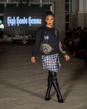Runway model wearing plaid skirt with black sweater and tall boots at fashion show.